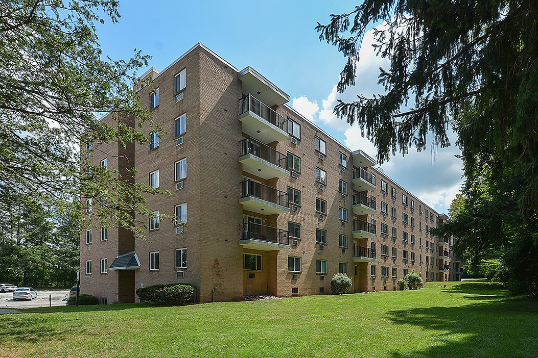 Exterior of an apartment building in East Norriton, PA with a large grassy area, large trees, and parking spots.