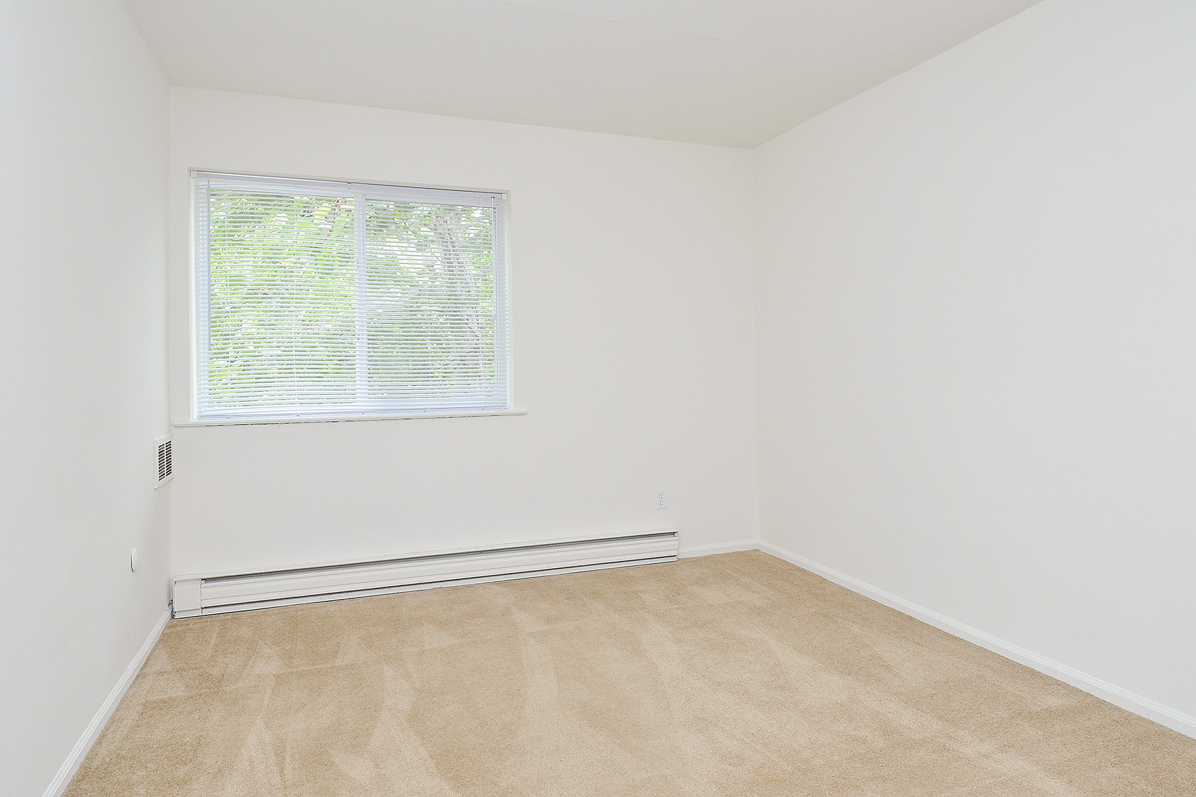 Spacious bedroom with a large window, white walls, and beige carpeting.