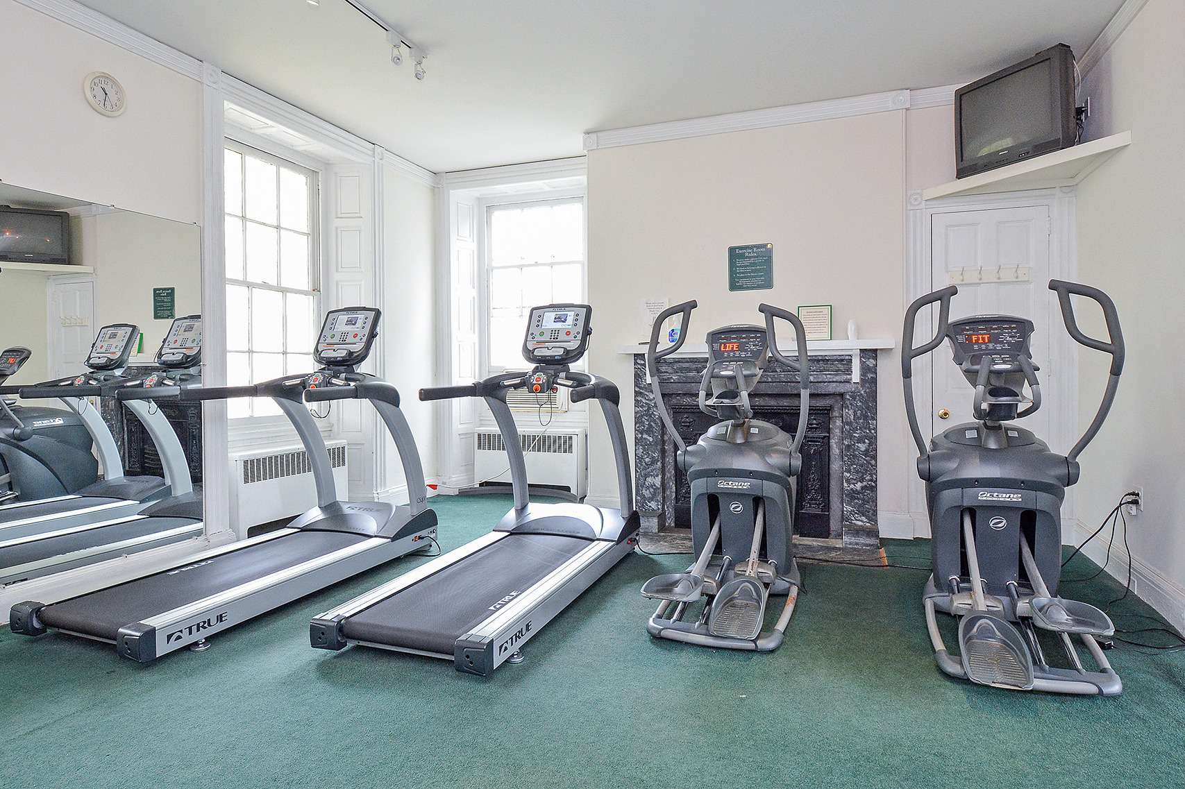 A variety of workout equipment in front of two windows, a mirror, and a tv in the top corner of an indoor gym.