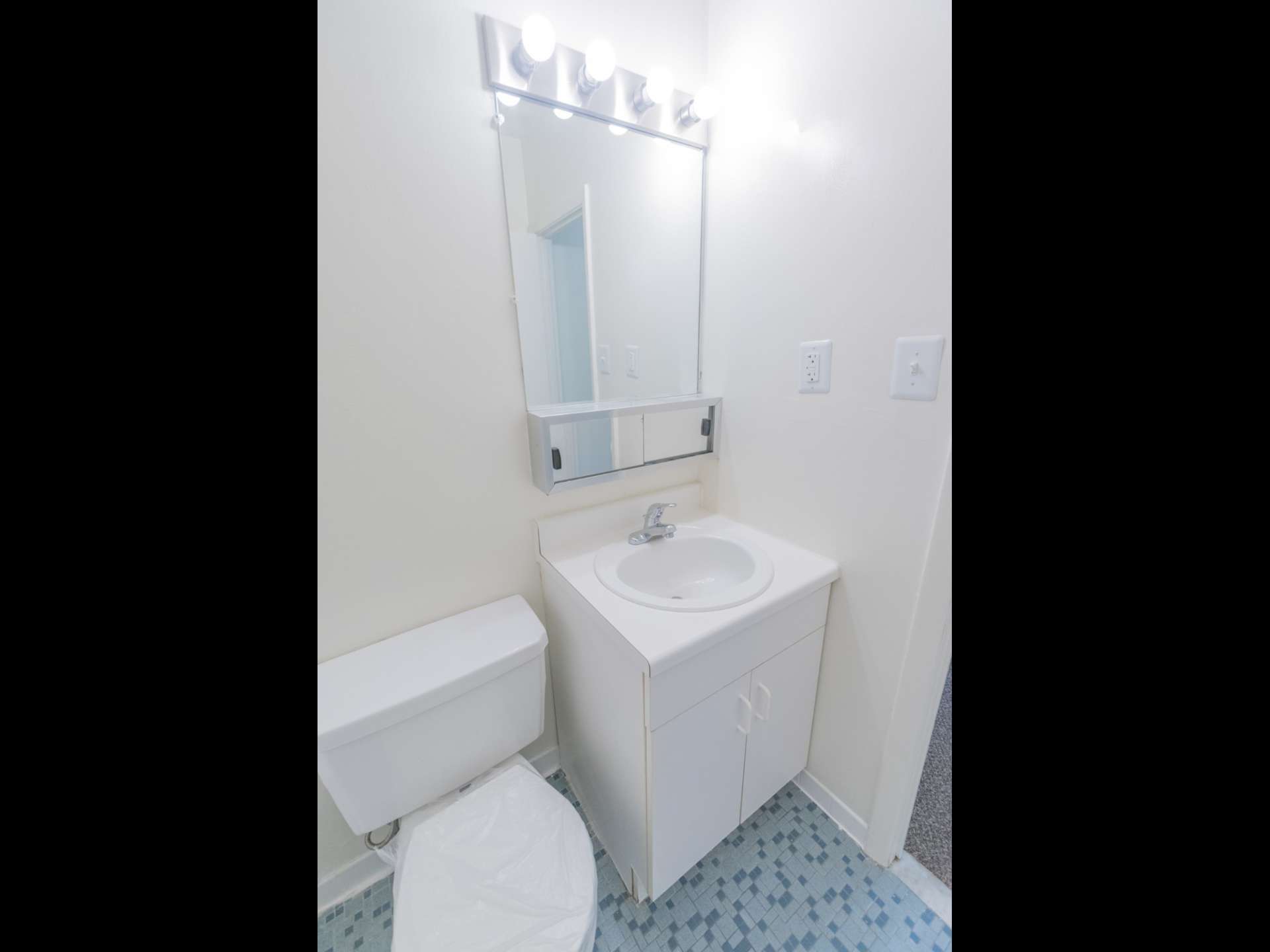 Bathroom with blue tile flooring, mirror, toilet, and white sink with cabinets.