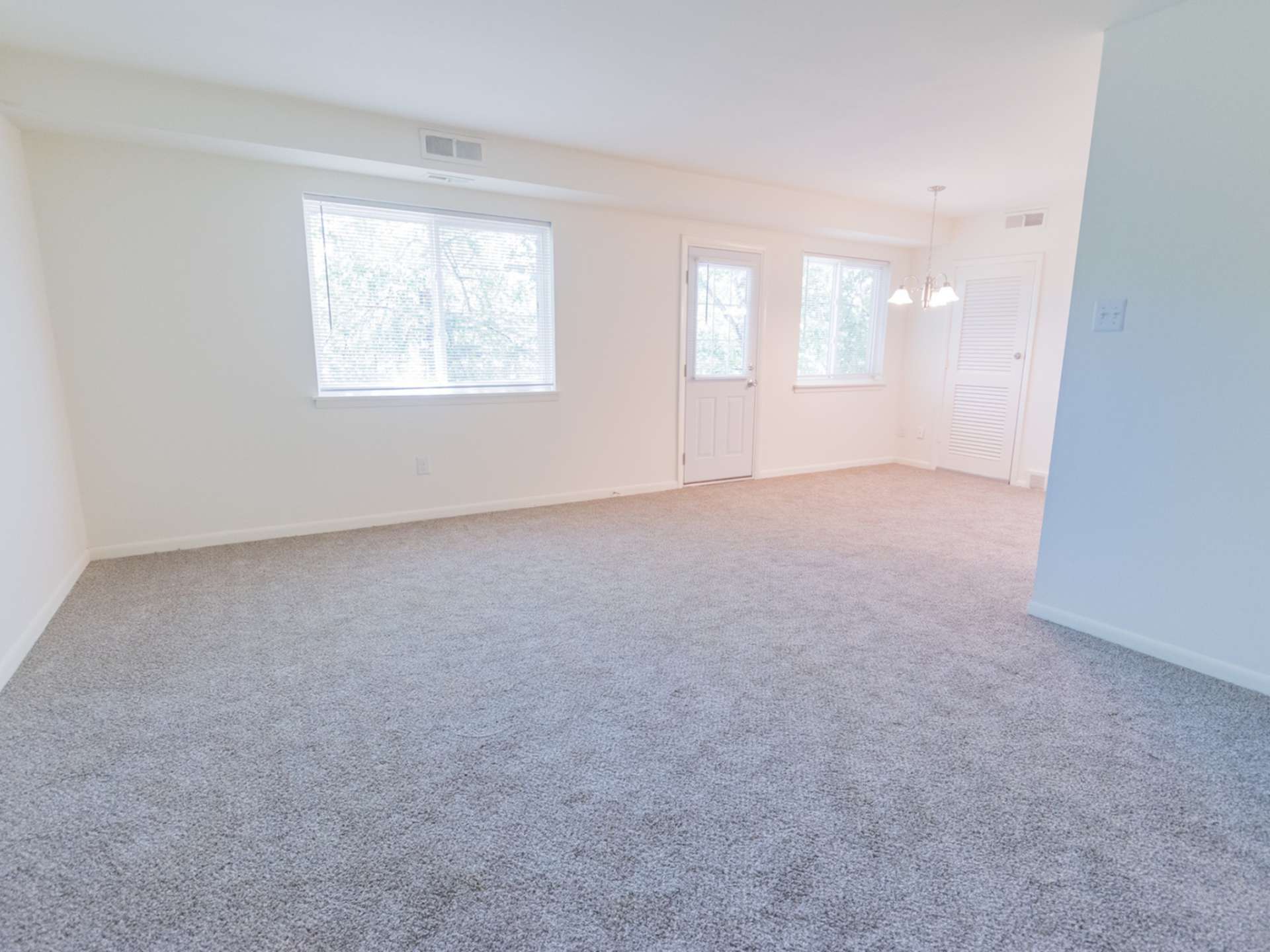 Spacious living area with a hanging light, two large windows, and light grey carpeting.