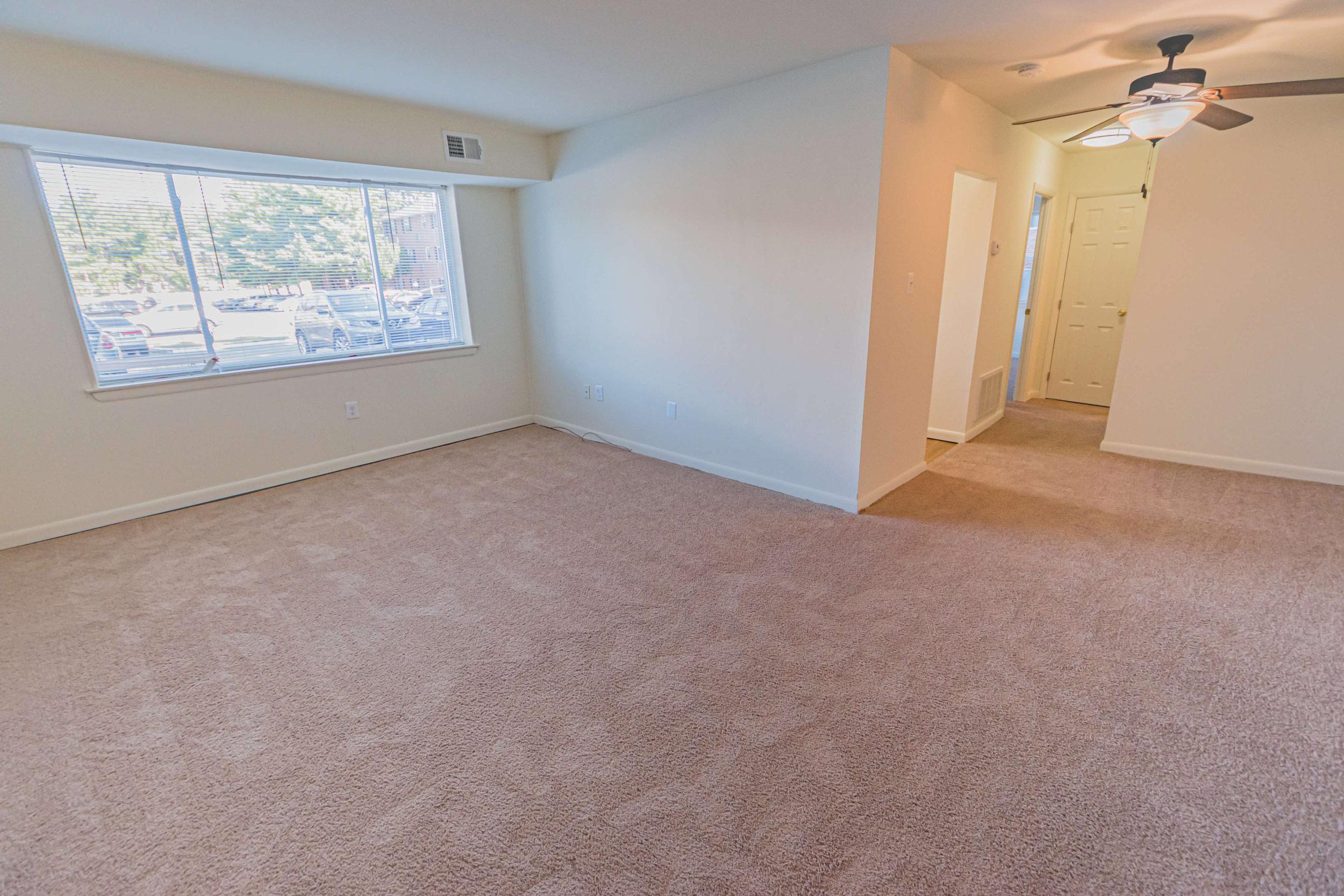 The living room area of an apartment, fitted with carpet flooring, a closet, and a huge window