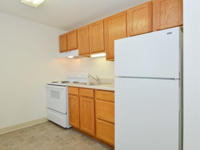 Kitchen area of an apartment, fitted with tiled flooring, wooden cabinets, a stove, and a fridge
