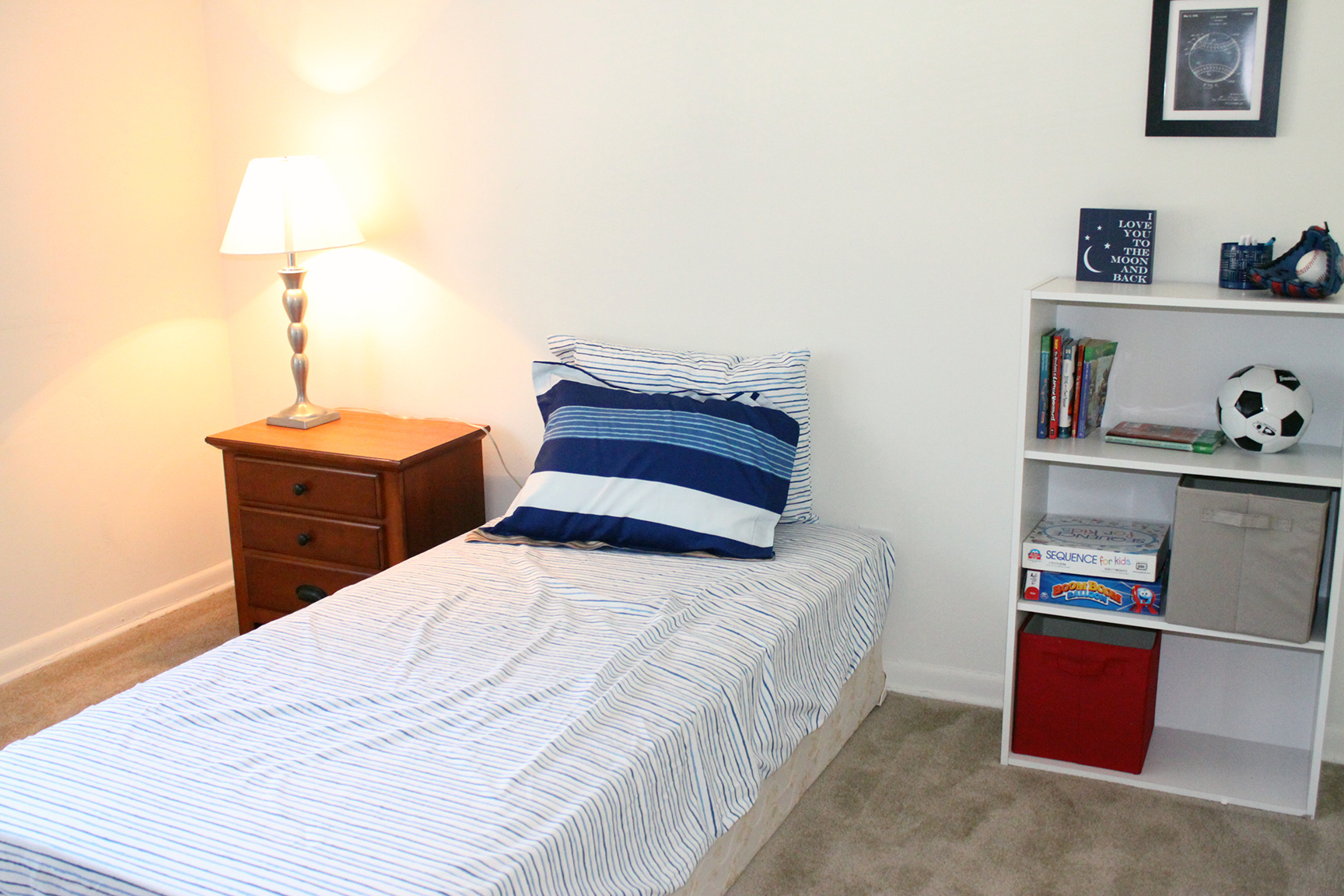 Bedroom area of an apartment, fitted with a twin size bed, carpet flooring, and a night stand