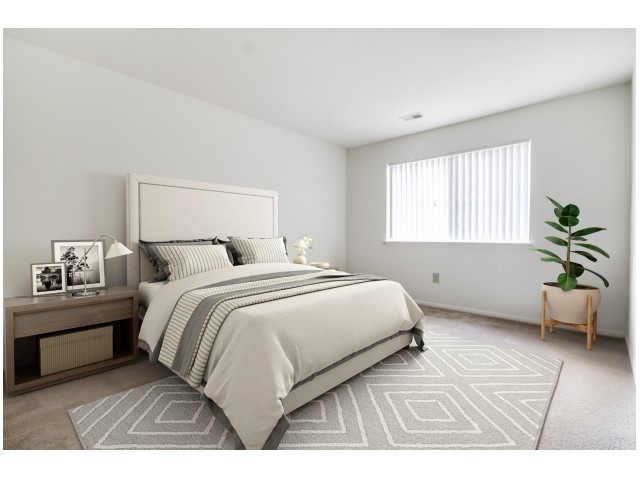 Furnished bedroom with light grey carpets and a large window at Indian Run Apartments.