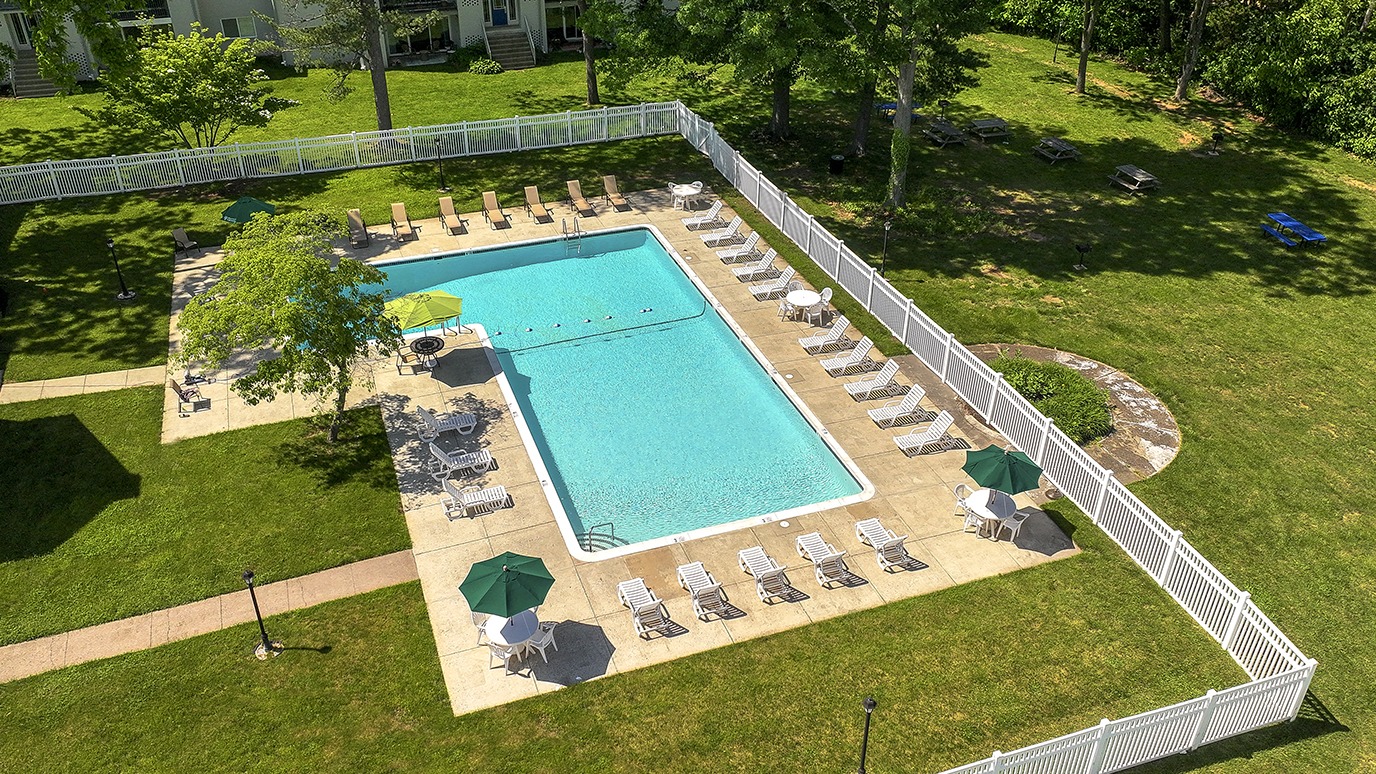 Drone view of an L-shaped gated swimming pool in a paved area surrounded by a large lawn